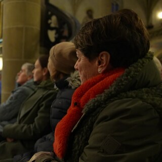 Sonja Faber-Schrecklein sitzt in einer Kirche auf einer Bank. Sie schaut zu einer kleinen Bühne, auf der ein Konzert gegeben wird. 