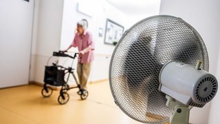 Eine Frau geht mit einem Rollator in einem Seniorenheim über einen Flur, der aufgrund hoher Außentemperaturen mit einem Ventilator belüftet wird.
