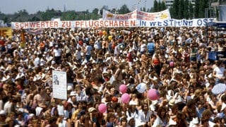 Demonstration auf den Rheinwiesen bei Bonn gegen die nukleare Nachrüstung am 10. Juni 1982. Etwa 500.000 Menschen nahmen teil. Im Hintergrund ein großes Transparent mit der Aufschrift: "Schwerter zu Pflugscharen – In West und Ost". Das Zitat "Schwerter zu Pflugscharen" hat seinen Ursprung in der Bibel und wurde auch in der Friedensbewegung zum geflügelten Wort.
