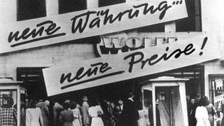 Kundenandrang vor einem Geschäft, das nach der Währungsreform 1948 Schilder mit der Aufschrift "Neue Währung... neue Preise!" an der Hausfassade angebracht hat.