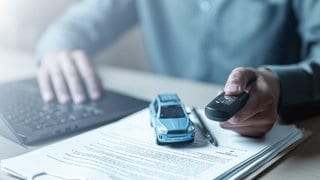 Zu sehen sind die Hände einer Person, die einen Autoschlüssel hält und mit der anderen Hand am Computer tippt. Die Person sitzt an einem Tisch. Darauf liegen auch Papierdokumente.