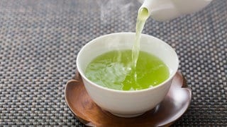 Grüner, dampfender Tee wird aus weißer Porzellankanne in weiße Tasse gegossen. Wie gesund sind grüner Tee und Matcha tatsächlich?