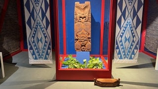 Maori-Holzskulptur aus dem 18. Jahrhundert in einer Vitrine auf Schloss Hohentübingen, flankiert von gemusterten Stoffbahnen.