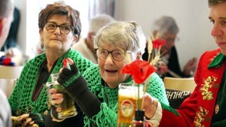 Mit fast 91 Jahren ist Thea Breuling als Lausbühlhexe bei der Fasnet in Ergenzingen aktiv. Sie sitzt mit anderen Narren in einem Saal, trägt einen grünen Umhang und trinkt ein Bier.