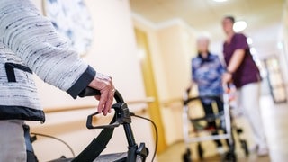 Senioren mit Rollator und eine Pflegekraft sind auf einem Gang in einem Pflegeheim zu sehen (Symbolbild).
