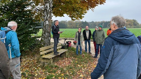 Martin Ruff vom Ulmer Klimanetz steht auf einer Bank neben einem Baum. Vor ihm einige der Personen, an dem Spaziergang durch das Waldgebiet Großer Gehrn bei Ulm-Jungingen teilgenommen haben, in dem vielleicht bald Windräder gebaut werden. 