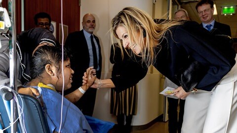 Eine blonde langhaaringe Frau, Königin Máxima, beugt sich zu einer Patientin im Rollstuhl, lächelt sie an und drückt ihre Hand.