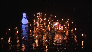 In einem Fluss schwimmen Menschen in Neoprenanzügen. Jeder hält eine Fackel über der Wasseroberfläche. Ein mit Luft gefüllter, schwimmender Schneemann treibt auf dem Fluss.  