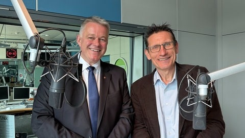 Ministerpräsident Gordon Schnieder und Korrespondent Chris Berdow stehen im SWR-Studio in Mainz vor einem Stehtisch mit Mikrofonen nebeneinander und schauen in die Kamera