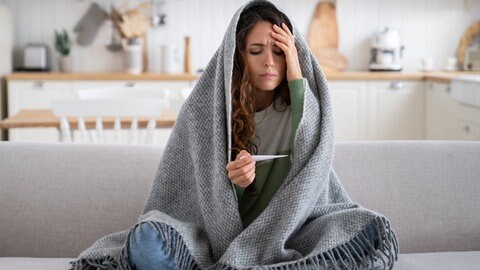 Frau sitzt unter einer Decke, hält sich den Kopf und hat Thermometer in der Hand.