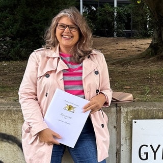 Susanne Sarantis hat mit 56 Jahren Abitur gemacht. Sie steht vor der Schule und hält ihr Abiturzeugnis. Neben ihr ist ein Schild mit dem Wort Abendgymnasium.