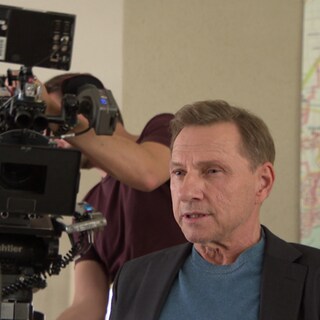 Richy Müller sitzt am Filmset, hinter ihm ist eine professionelle Kamera für die Dreharbeiten zu sehen.
