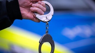 Ein Polizist vor einem Polizeiauto hält ein Paar Handschellen (Symbolbild): In Wörth am Rhein ist ein Mann nach einer Schlägerei im Bahnhof festgenommen worden.