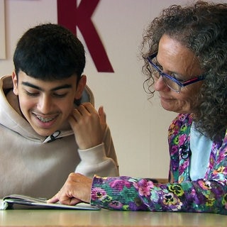 Der 15-jährige Abdul und seine Mentorin Karin Zahn sitzen an einem Tisch und schauen gemeinsam auf ein Schulbuch.