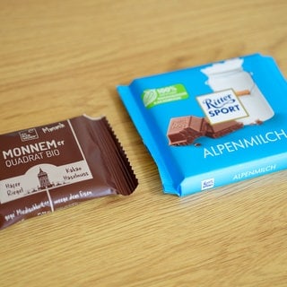 Eine Tafel mit Schokolade der Marke Ritter Sport (r) und ein Haferriegel der Firma Wacker mit der Aufschrift "Monnemer Quadrat Bio" liegen nebeneinander auf einem Tisch. 
