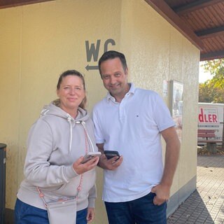 Maurice Skubski und seine Schwägerin Bianca Kauffmann stehen vor einer öffentlichen Toilette in Renningen und halten ihr Smartphone in der Hand.