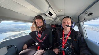 Marleen und ihr Verlobter Matthias sitzen im Flugzeug. Durch die Fenster ist der Bodensee zu sehen.