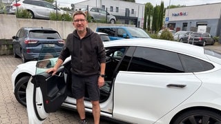 Jochen Melcher steht auf einem Hof hinter der offenen Tür seines weißen Tesla Model S