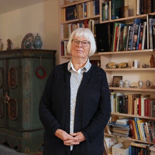 Zeitzeugin Mechthild wird kurz nach dem Zweiten Weltkrieg geboren. Sie steht bei sich zu Hause im Wohnzimmer vor einem Bücherregal. Sie hat weiße Haare, trägt eine blaue Brille und schaut in die Kamera.