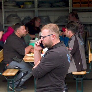 Ein Mann im Vordergrund trägt Arbeitskleidung und telefoniert. Im Hintergrund sitzen mehrere Menschen auf Bierbänken und essen.