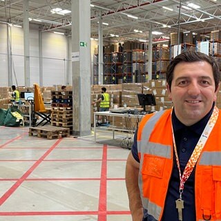 Logistiker Kreshnik Himaj steht lächelnd in einer Lagerhalle vor gestapelten Paketen.