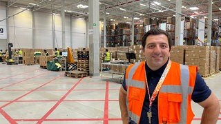 Logistiker Kreshnik Himaj steht lächelnd in einer Lagerhalle vor gestapelten Paketen.