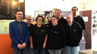 Eine Gruppe von sieben Menschen steht vor einer Café-Theke. Alle tragen schwarze Westen mit der roten Aufschrift „Ella’s“. Der Mann ganz links trägt stattdessen einen dunkelblauen Anzug.