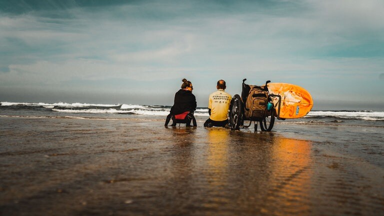 Mit dem Rücken zur Kamera: Der querschnittgelähmte Johannes Laing sitzt mit einer gleichaltrigen Frau am Strand und sie beobachten die Wellen. 