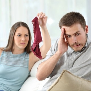 Erwischt! Wütende junge Frau präsentiert ihrem beschämten Partner fremde rote Damenunterwäsche: Treue gilt als Basis für eine erfüllte Liebesbeziehung. Ein Seitensprung ist daher die häufigste Ursache für Konflikte und Trennungen.