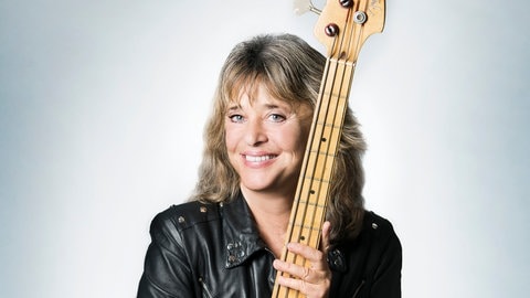 Sängerin Suzi Quatro steht in einer Lederjacke und einer Gitarre in der Hand und schaut direkt in die Kamera