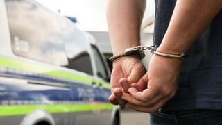 Ein Mann in Handschellen neben einem Polizeifahrzeug: Ein Verkehrsunfall vor einem Kindergarten in Waldenburg entpuppte sich als versuchte Tötung. Die Polizei hat drei Verdächtige festgenommen.