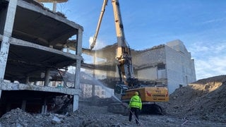 Ein Abrisskran bei der Arbeit. Daneben steht ein Arbeiter und spritzt Wasser, um den aufgewirbelten Staub zu reduzieren. Der Abriss des Rathaus-Centers Ludwigshafen schreitet voran.