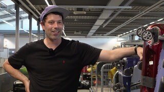 Michael Höll steht im Kofferlager des Flughafens Baden-BadenKarlsruhe und stützt sich auf einen Stapel Reisekoffer ab. Er trägt eine lila Mütze und ein dunkles T-Shirt.