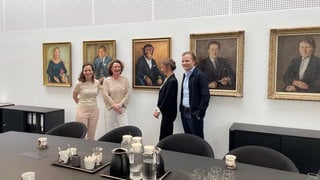 Die Grupp-Geschäftsführer stehen mit den Künstlerinnen lachend vor der Ahnengalerie im Trigema-Firmengebäude. Über ihnen zwischen Ahnenbildern das neue Gemälde mit Affe Charly. 