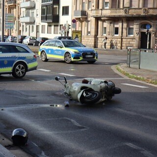 Auf der Straße liegt ein Roller auf der Seite. Davor liegt ein Helm. Hinter dem Auto sieht man zwei Polizeiautos, die die Straße absperren.