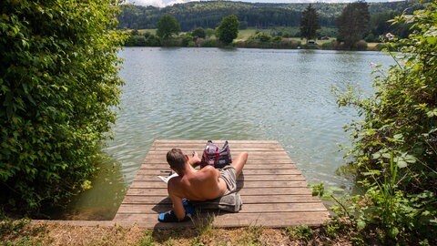 Ein Mann liegt auf dem Steg eines Badesees. In Rheinland-Pfalz wird es in den kommenden Tagen spätsommerlich warm.