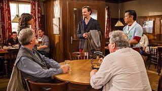 Schorsch und Leo sitzen am Stammtisch, an der Tür stehen Tu und Eva und schauen auf einen neuen Gast