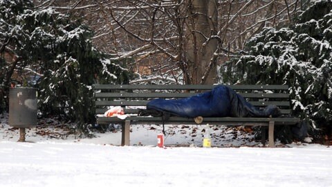 Ein Obdachloser liegt im Schlafsack auf einer Parkbank im Schnee.