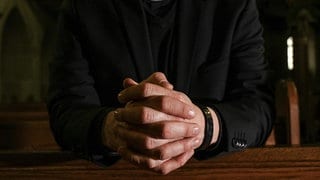 Das Symbolfoto zeigt einen Priester, er kniet in einer Kirche und hat die Hände fest zum Gebet gefaltet. In Koblenz beginnt das Berufungsverfahren gegen einen umstrittenen katholischen Priester aus dem Westerwald, dem Kindesmissbrauch vorgeworfen wird.