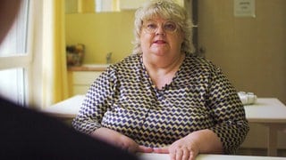 Eine Frau mittleren Alters mit kurzen, blonden Haaren und runder Brille spricht an der Kamera vorbei zu einer Person.