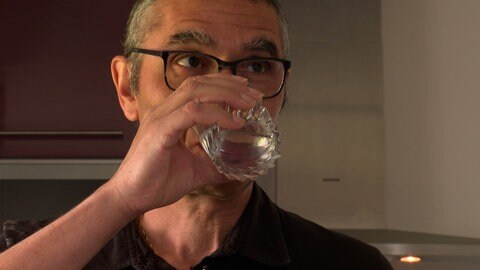 Mann trägt eine Brille und weiße Haare. Er trinkt aus einem Wasserglas. Man sieht nur seine Augen.