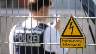 Ein Polizist steht in einem Umspannwerk an einem Zaun, an dem ein Schild mit der Aufschrift "Hochspannung Lebensgefahr" hängt. (Symbolbild)