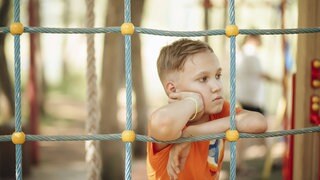 Ein traurig aussehender Junge lehnt sich auf einem Spielplatz in einer Kletternetz. 