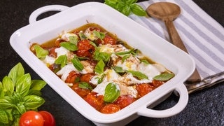 Rezept für Ofengericht: Fruchtige Tomaten mit Burrata und Basilikum in einer Auflaufform. Kochlöffel auf gestreiftem Küchentuch liegt daneben. Auf der anderen Seite liegen Tomaten und Basilikumblätter.