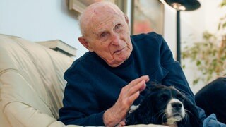 Josef kümmert sich dreimal die Woche um Leihhündin Elli. Der 104-Jährige sitzt mit Elli auf einem beigen Sofa. Josef streichelt Elli und sie kuscheln gemeinsam. 