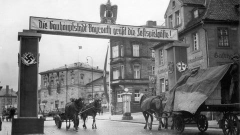 Historische Aufnahme eines Straßenschilds in Bayreuth aus dem Jahr 1944 mit der Aufschrift „Die Gauhauptstadt Bayreuth grüßt die Festspielgäste“. Über dem Schild ist ein Reichsadler angebracht. Die Aufnahme stammt aus der Zeit der NS-Herrschaft und der sogenannten Kriegsfestspiele.