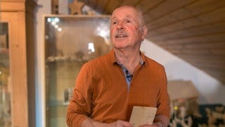 Armin steht vor einer Wand aus Holz, wo auch einige seiner Schnitzereien zu sehen sind. Er hat ein Holzbrett in der Hand und schaut neben der Kamera vorbei. 