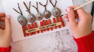 Wundervolles Winterwissen - der SWR1 Adventskalender: Warum macht uns basteln im Advent und an Weihnachten glücklich? Das Bild zeigt zwei Frauenhände, die Wäscheklammern auf einer Weihnachtskarte mit roter Farbe bemalt. Sie sind zusammen mit kleinen Steinen und Zweigen auf der Karte aufgeklebt - es sieht so aus, als schauten Rentiere über einen Zaun. Ein Schriftzug sagt "Merry Christmas"
