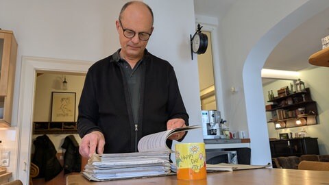 Thomas Mann blättert seine Bescheide zur Grundsteuer durch. Er freut sich über eine niedreigere Grundsteuer. Vor ihm Steht eine Tasse, auf der Happy Day steht.
