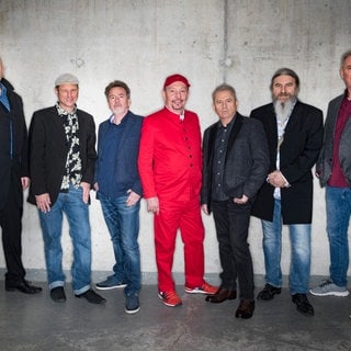 Die Spider Murphy Gang steht in einer Reihe vor einer Wand und alle schauen in die Kamera.